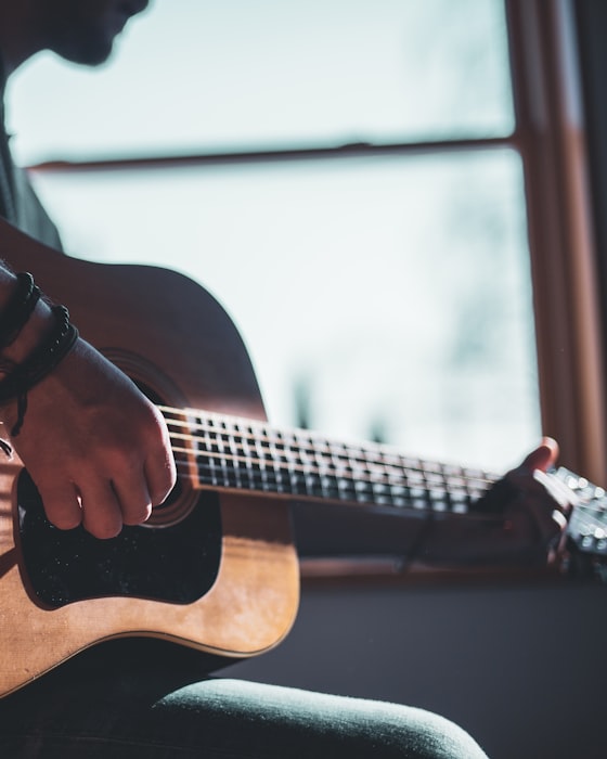 A guitarist playing a steel-string acoustic guitar in warm window light